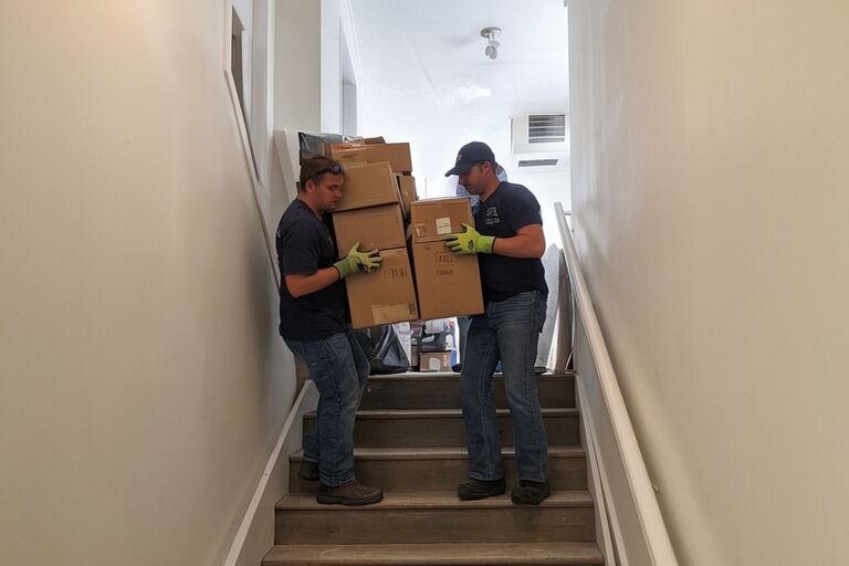 Workers carrying stacked boxes up steep basement stairs in a Staten Island home