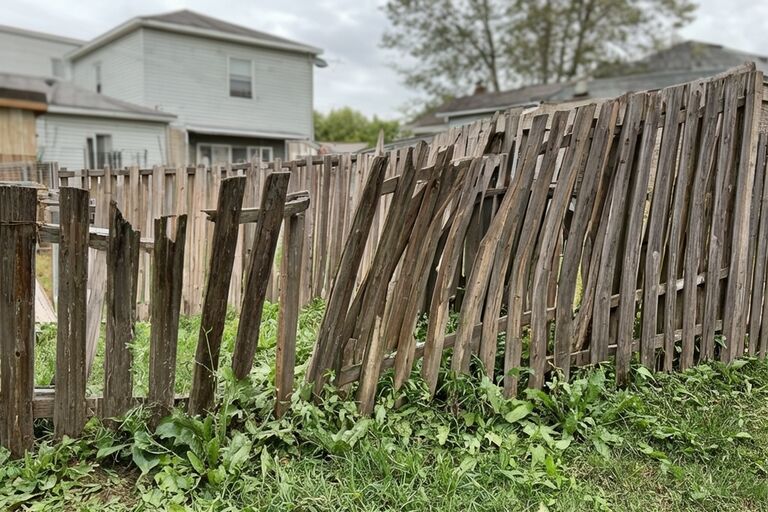 Old leaning wooden fence with broken pickets and tilting posts in residential yard